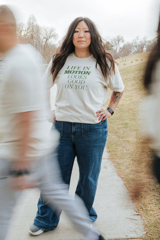 Woman wearing a white t-shirt with text that says "Life in motion looks good on you", standing outdoors on a path.