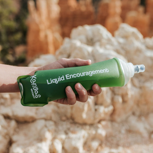 Person holding a green soft flask with 'Liquid Encouragements' text against a natural background