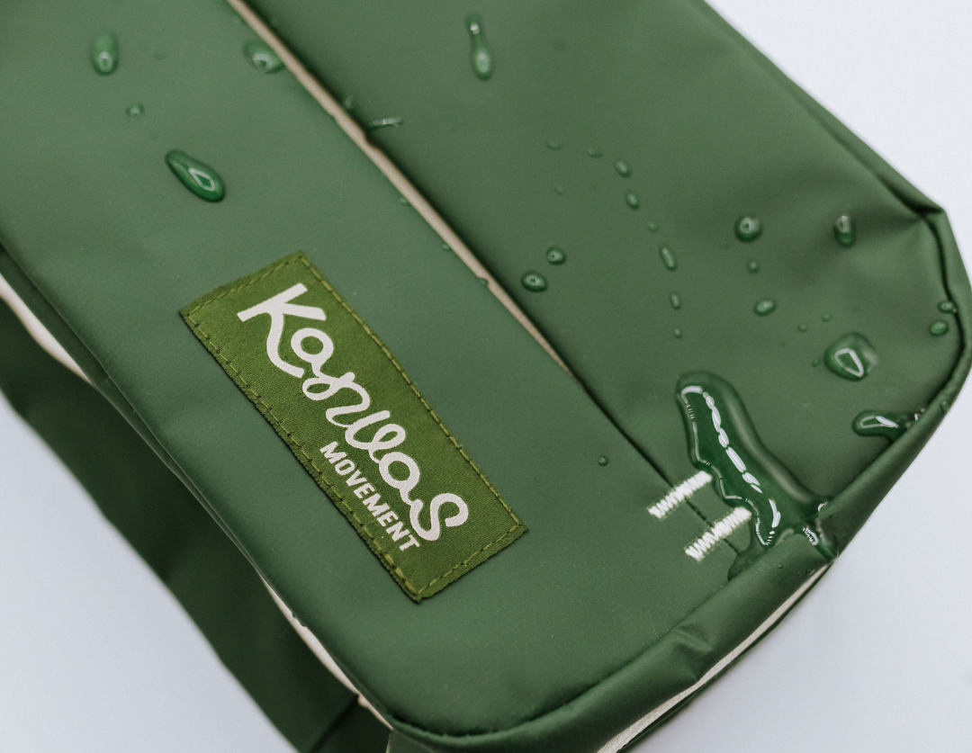 A dark green waterproof pouch with a visible Kanvas Movement logo patch, shown with water droplets on the surface. 