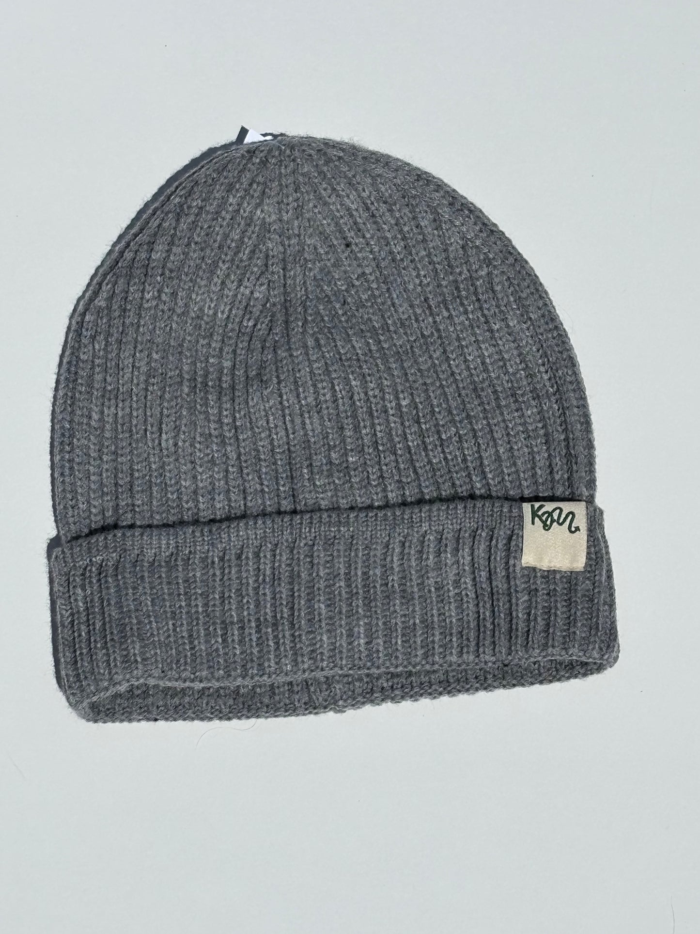 A ribbed knit beanie hat in a light grey color, featuring a small white fabric brand patch on the folded cuff with the text 'KM'.