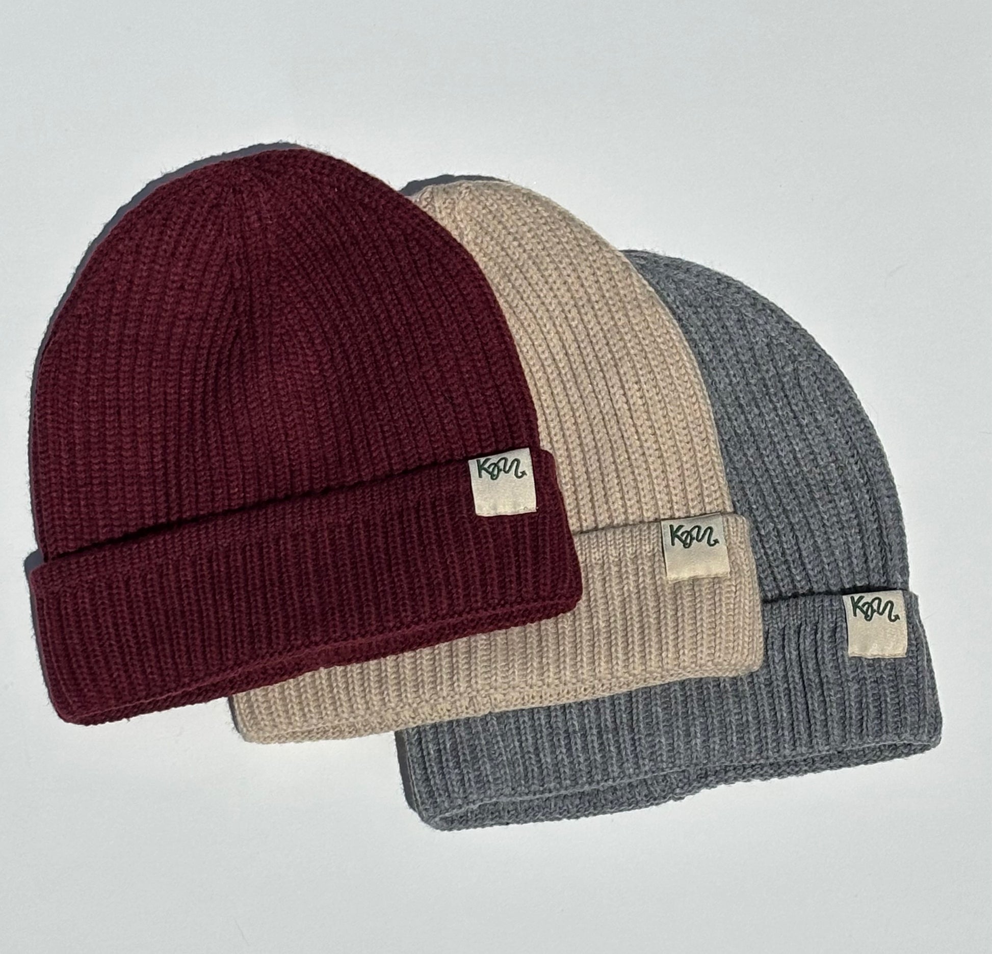 Three ribbed knit beanie hats in burgundy, beige, and heather gray, arranged in a fan layout on a light gray background. Each beanie has a small rectangular fabric tag on the folded cuff with the brand name 'KM' printed on it.