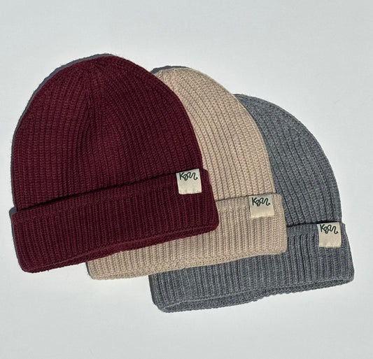Three ribbed knit beanie hats in burgundy, beige, and heather gray, arranged in a fan layout on a light gray background. Each beanie has a small rectangular fabric tag on the folded cuff with the brand name 'KM' printed on it.