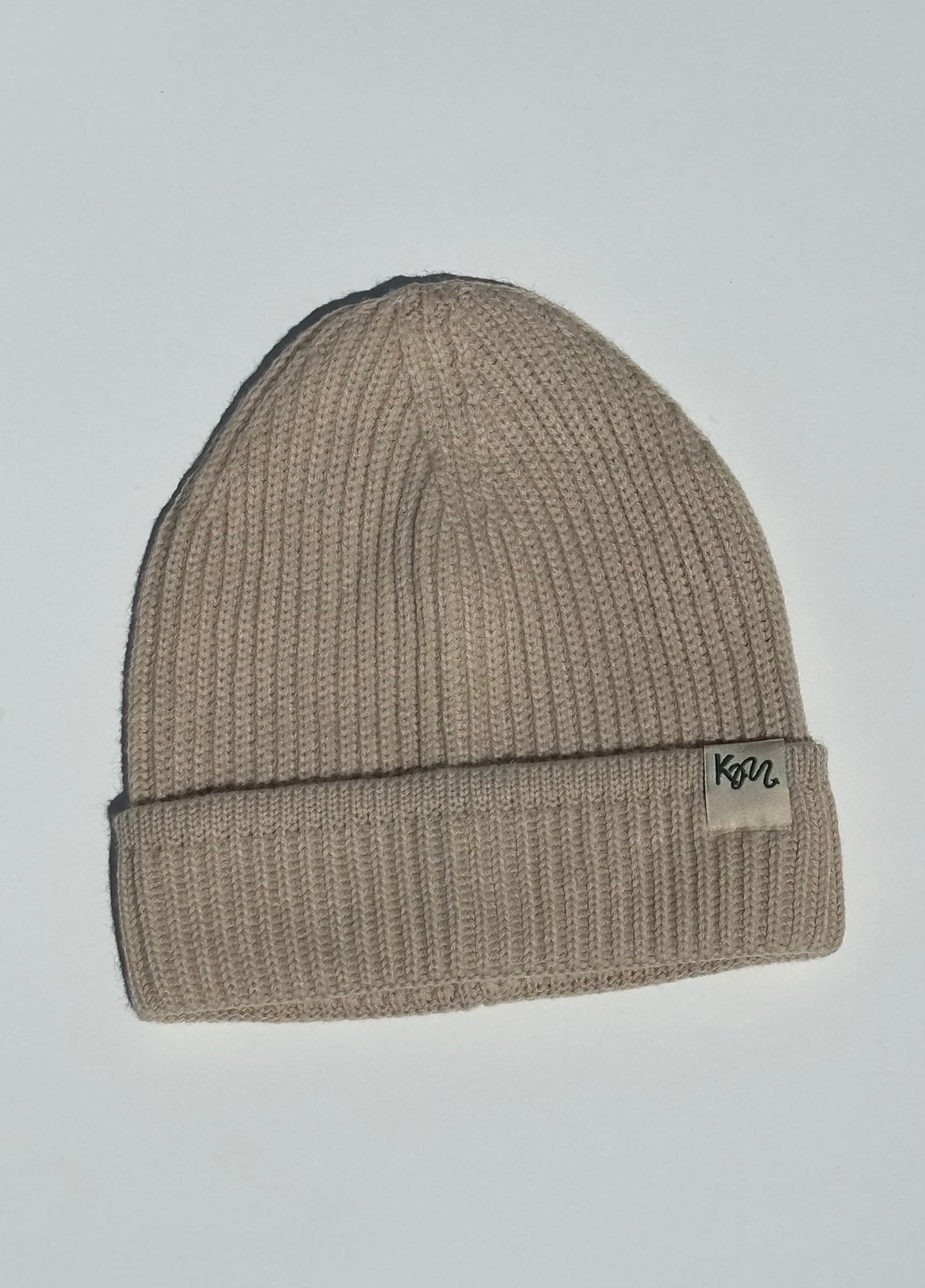 A ribbed knit beanie hat in a light taupe-beige color, featuring a small white fabric brand patch on the folded cuff with the text 'KM'.