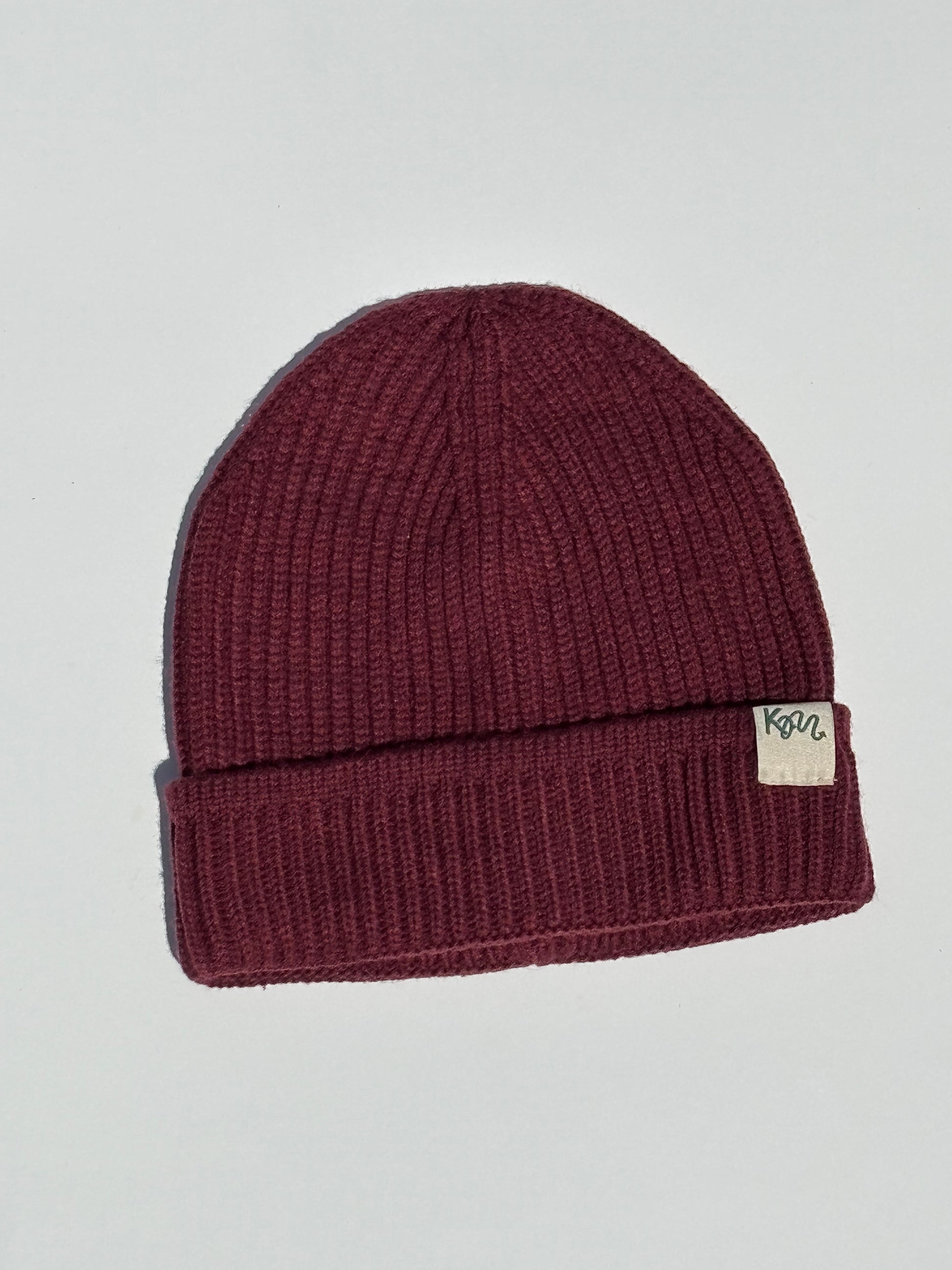A ribbed knit beanie hat in a deep burgundy or maroon color, featuring a small beige fabric brand patch that says 'KM' on the folded cuff. The beanie is photographed flat against a light gray background.