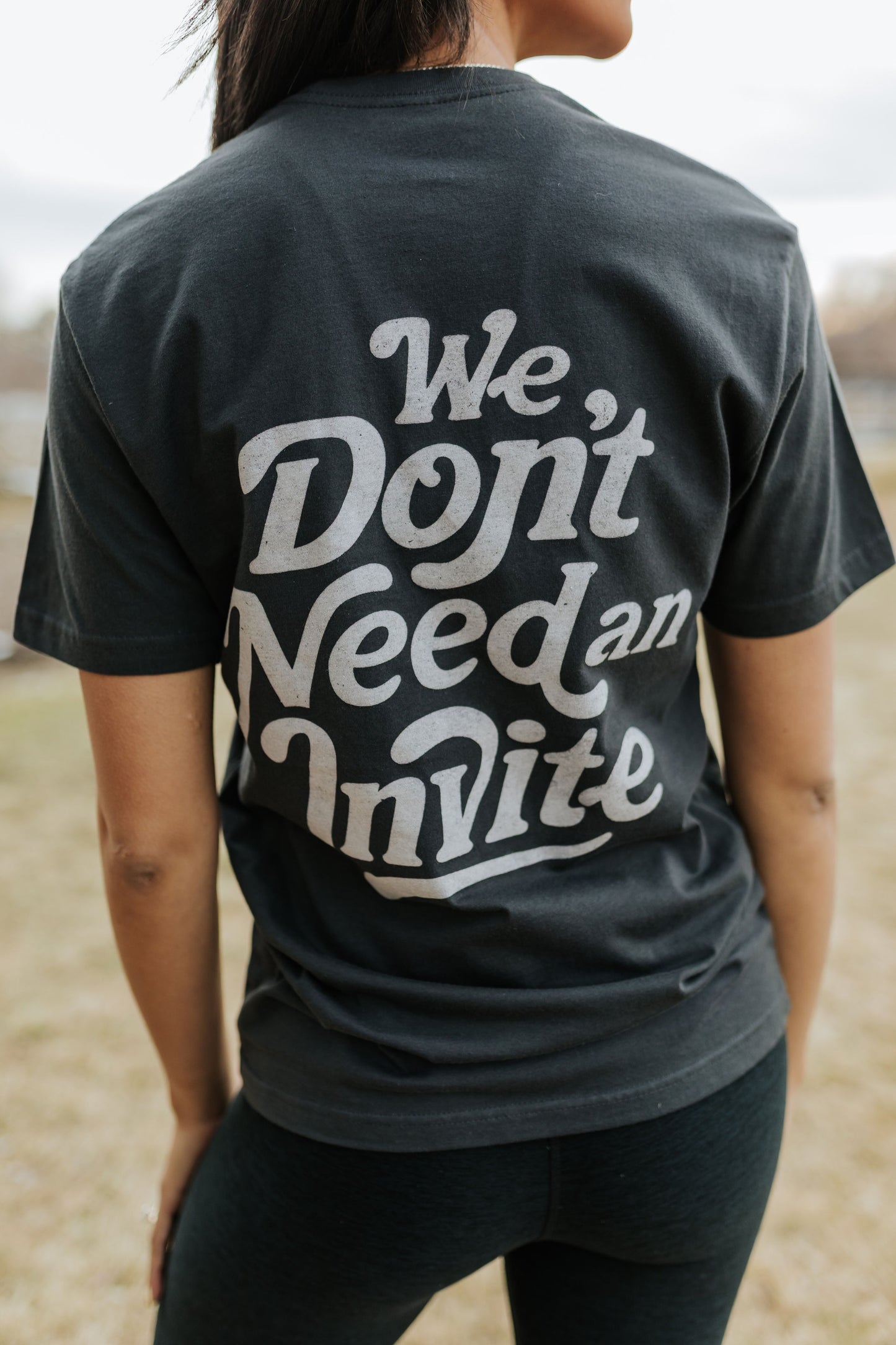 Person wearing a black t-shirt with 'We Don't Need an Invite' text in an outdoor setting.
